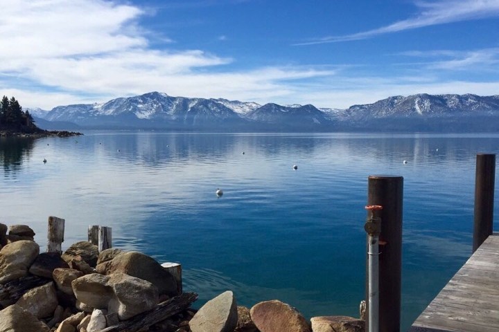 Serene lake with rocky shore, distant snowy mountains, and blue sky.