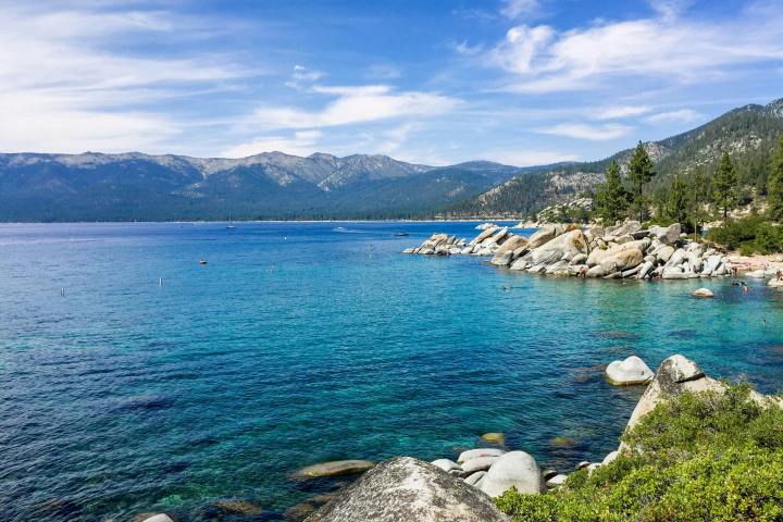 Clear blue lake with rocky shoreline and mountains under a partly cloudy sky.