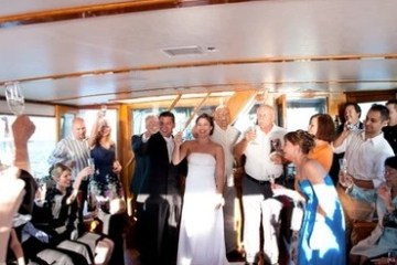 Group celebrating on a boat, people raising glasses, bride in white dress.