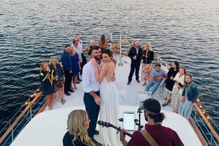 Couple in wedding attire dance on a boat surrounded by guests and musicians.